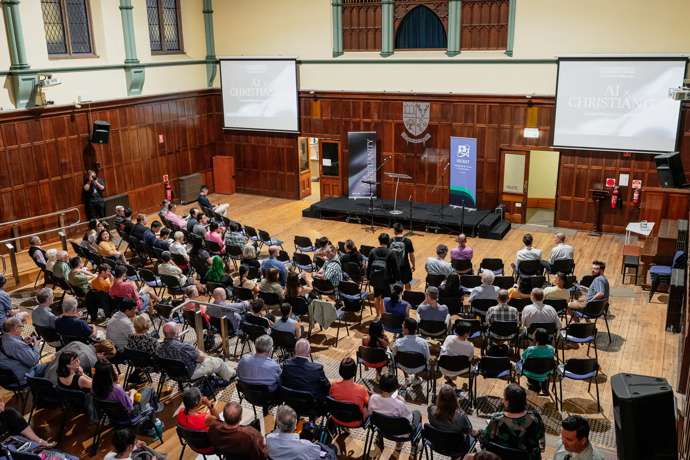 An audience seated in Werner Brodbeck Hall, with timber-panelled walls and a stage at the front